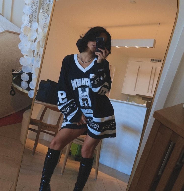 Becky G com corte bob está em frente a um espelho, vestindo uma camiseta oversized preta com estampas brancas de estilo esportivo. Ela usa botas de cano alto pretas e segura o celular para tirar a foto. O ambiente é uma sala bem iluminada. 