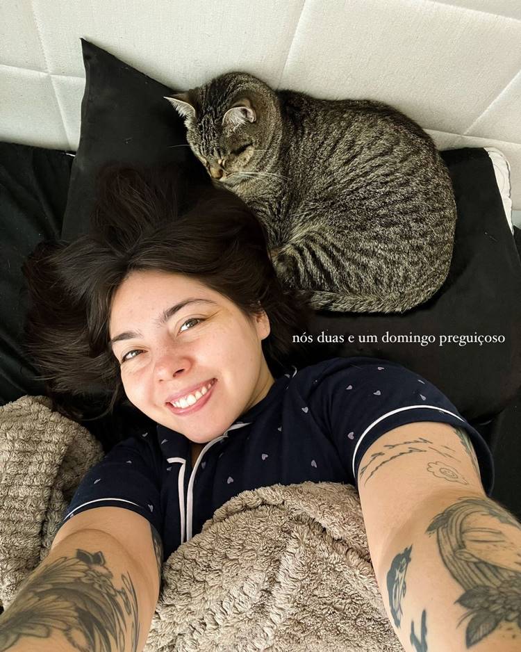 Mulher deitada sorrindo ao lado de um gato cinza dormindo em um travesseiro preto. Ela usa pijama azul com detalhes brancos e tem tatuagens nos braços. Estão cobertos por um cobertor felpudo, transmitindo uma sensação de conforto e preguiça. Ela é uma das meninas da lista de blogueiros CLTs.