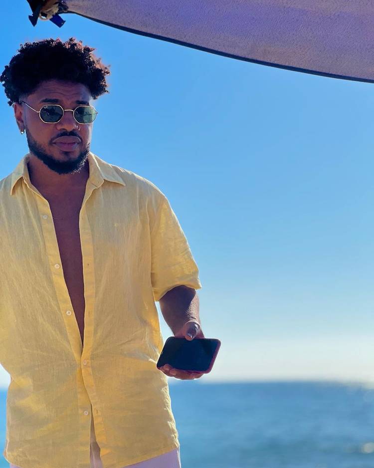 Homem de óculos escuros e barba usa camisa amarela aberta, segurando um celular. Ele está em uma praia sob um guarda-sol, com o mar azul ao fundo e céu claro, transmitindo uma sensação de lazer e tranquilidade.