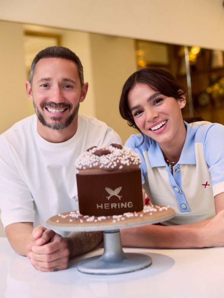 Cedric Grolet usando blusa branca e Bruna Marquezine usando blusa azul e bege de gola, ambos apoiados sobre mesa com panetone de chocolate embalado com logo da Hering sobre boleira ao centro