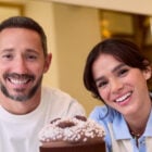 Cedric Grolet usando blusa branca e Bruna Marquezine usando blusa azul e bege de gola, ambos apoiados sobre mesa com panetone de chocolate embalado sobre boleira ao centro