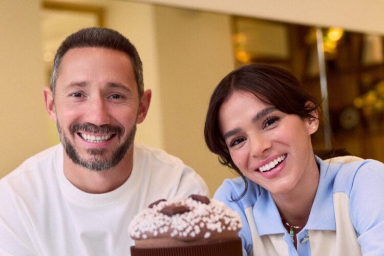 Cedric Grolet usando blusa branca e Bruna Marquezine usando blusa azul e bege de gola, ambos apoiados sobre mesa com panetone de chocolate embalado sobre boleira ao centro