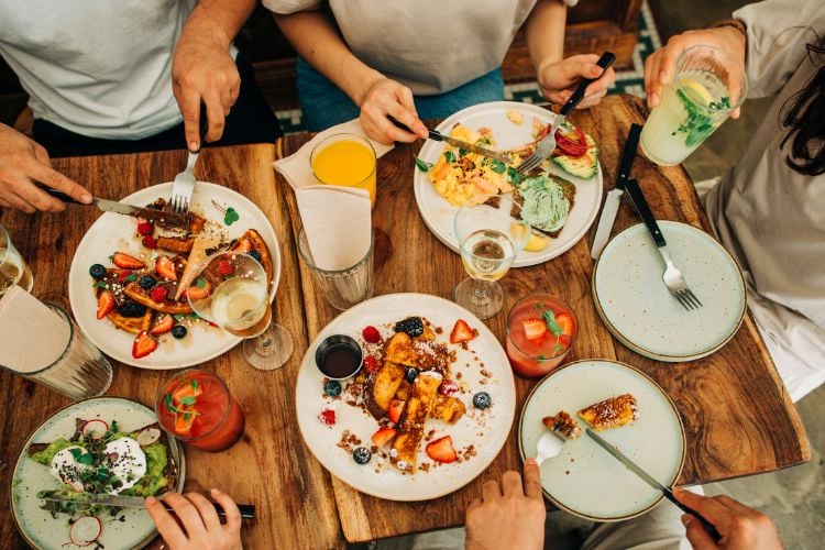 Pessoas de pele clara aparecendo apenas os braços, comendo comidas de brunch, como ovos mexidos e panquecas 