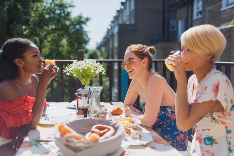 Três mulheres, duas de pele clara e uma de pele negra, tomando suco e rindo, sentadas à mesa com comidas típicas de brunch e arranjo de flores