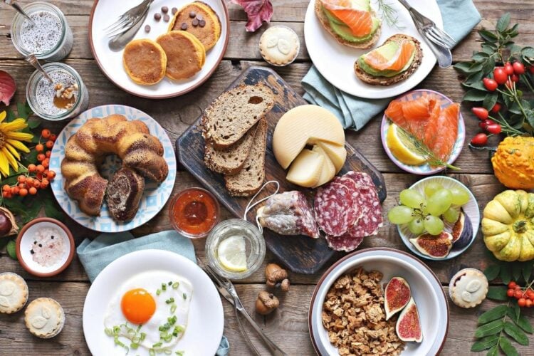 Mesa com bolo, pães, queijo, frios, frutas, panqueca, cereais, avocado toast e mais comidas de brunch