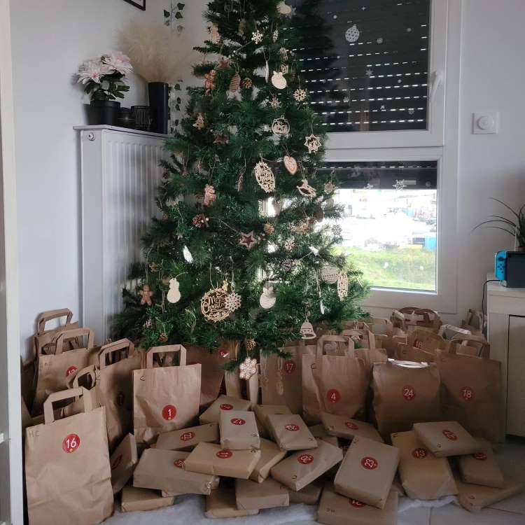 Árvore de Natal decorada com enfeites de madeira, cercada por sacolas de papel kraft numeradas, formando um calendário do advento. As sacolas estão dispostas de forma organizada sobre um pano branco. Ao fundo, uma janela e decoração simples completam o ambiente festivo.