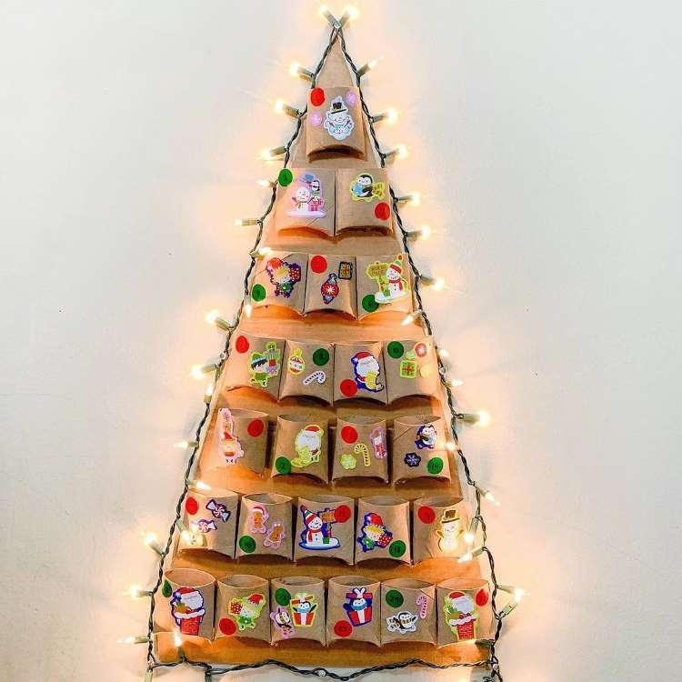 Um calendário do advento em forma de árvore é feito com envelopes de papel pardo decorados com adesivos natalinos. Luzes pisca-pisca contornam o formato triangular na parede, criando uma atmosfera festiva e encantadora, ideal para a contagem regressiva para o Natal.