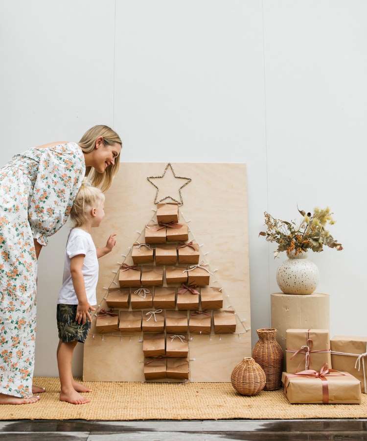 Uma mãe e seu filho admiram um calendário do advento em forma de árvore feito com sacos de papel pardo. A estrutura tem uma estrela no topo e está decorada com luzes. Ao lado, cestos e presentes complementam o cenário natalino.