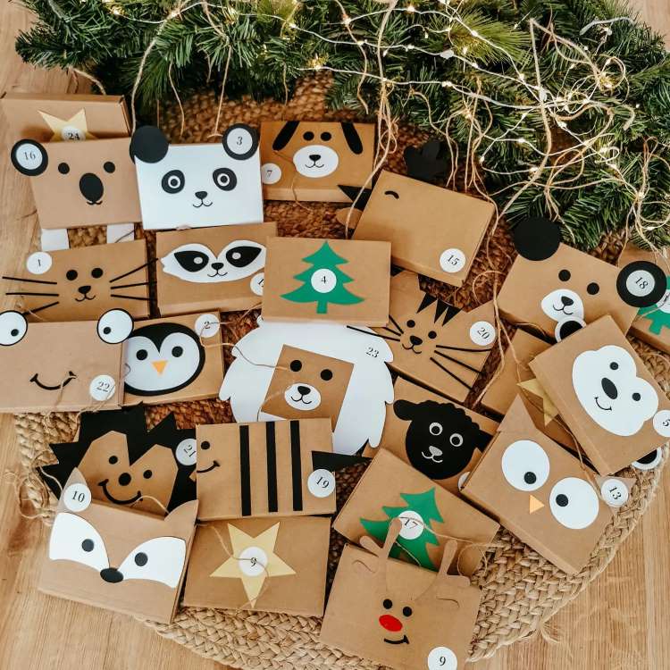 Calendário do advento feito com envelopes de papel pardo, cada um decorado como um animal diferente, incluindo panda, cachorro, pinguim e urso. Os envelopes estão numerados e dispostos sobre um tapete de juta, com galhos de pinheiro e luzes ao redor, criando um ambiente natalino encantador.