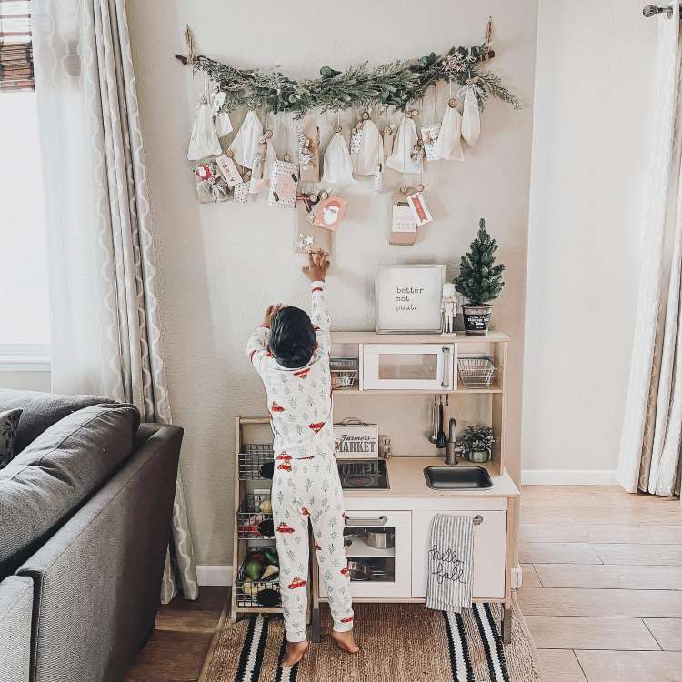 Criança em pijama decorado, alcançando um calendário do advento pendurado com sacolas e galhos de pinheiro. O ambiente é uma sala aconchegante, com cozinha de brinquedo ao fundo, cortinas claras e sofá, criando um clima natalino e acolhedor.