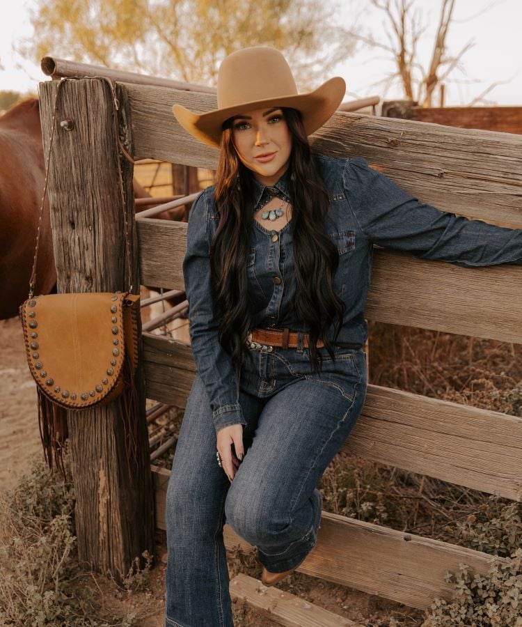 A imagem mostra uma mulher em um ambiente rústico, vestindo um conjunto jeans e um cinto marrom. Ela usa um chapéu de cowboy bege e colar com pingente de pedra azul. Sentada em uma cerca de madeira, ao lado de uma bolsa de couro com tachas, transmite um estilo country elegante.