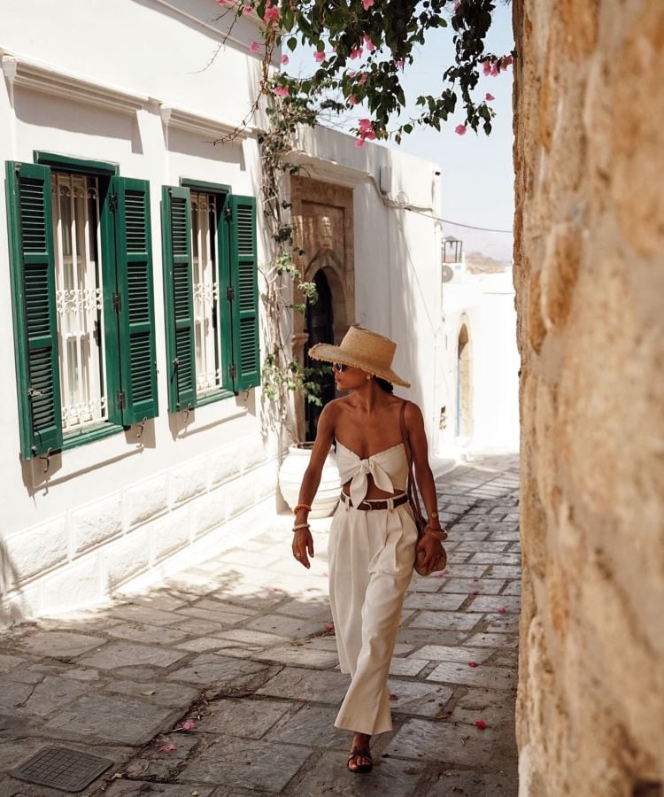 A imagem mostra uma mulher caminhando por uma rua pavimentada, cercada por paredes brancas com janelas de venezianas verdes. Ela usa um chapéu de palha, top branco com amarração, calça branca ampla e sandálias marrons. Ao fundo, flores rosas pendem de uma treliça, criando um cenário mediterrâneo ensolarado.