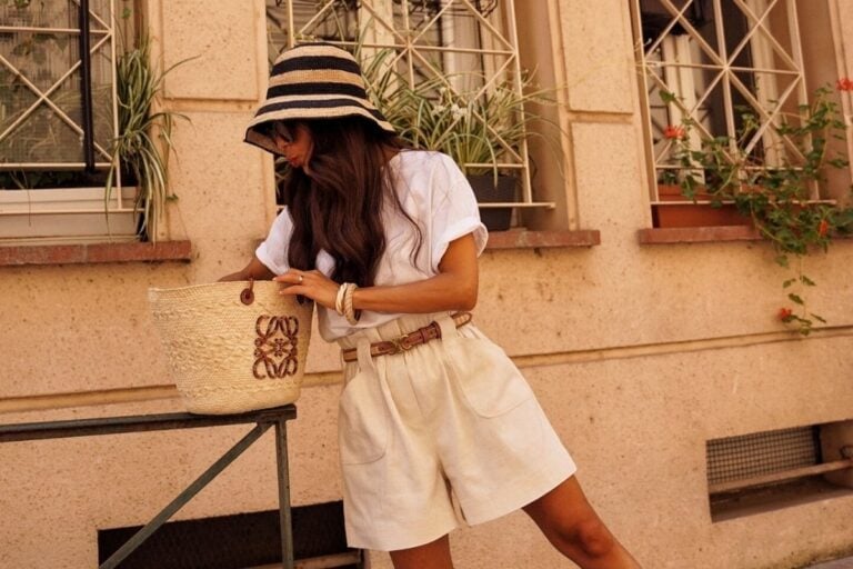 A imagem mostra uma mulher vestindo um chapéu listrado e uma blusa branca, combinados com shorts bege e um cinto marrom. Ela segura uma bolsa de palha com detalhes bordados. O cenário é urbano, com plantas em janelas ao fundo, criando um look casual e elegante.