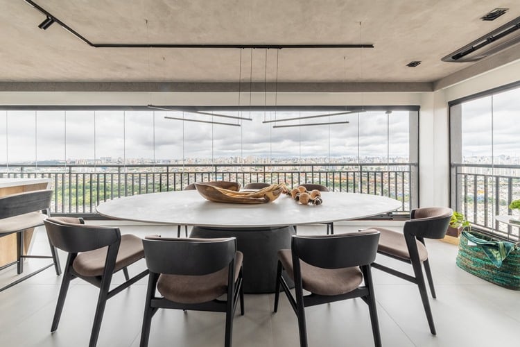 A foto mostra uma elegante sala de jantar com uma grande mesa redonda de madeira clara. Ao redor, há oito cadeiras acolchoadas em tons escuros. A sala é cercada por janelas amplas que oferecem uma vista panorâmica da cidade. O teto é moderno, com luminárias retangulares suspensas, e há objetos decorativos sobre a mesa.