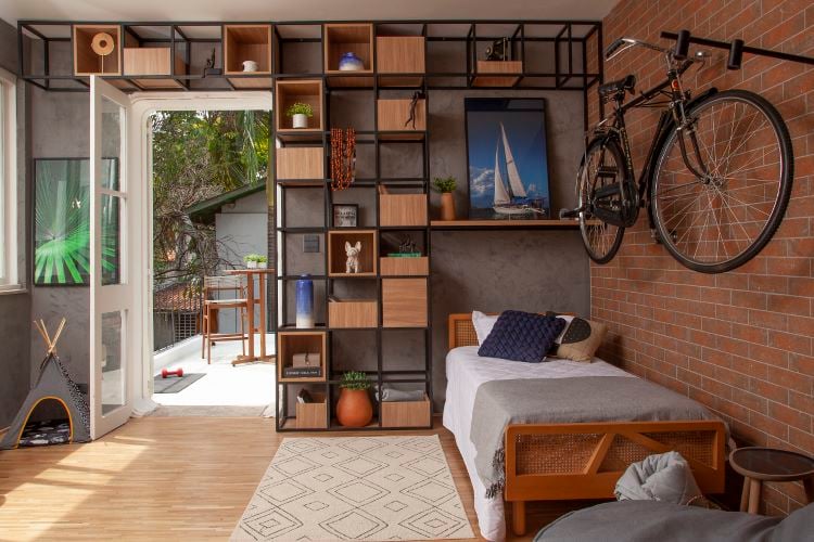 A foto mostra um quarto moderno e aconchegante com piso de madeira clara e uma parede de tijolos exposta. Há uma bicicleta pendurada na parede e uma estante com nichos decorativos ao lado de uma cama de solteiro com colchas cinza e travesseiros. A porta aberta revela uma varanda com plantas e móveis de madeira.