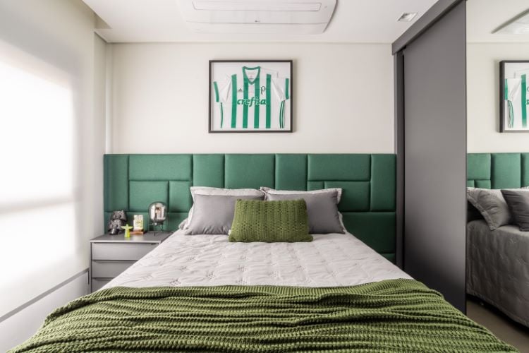 O quarto é moderno e bem iluminado, com uma cama de casal central. A cabeceira é acolchoada em verde, combinando com uma manta no pé da cama. Acima, há uma camisa de futebol emoldurada na parede. Ao lado da cama, um criado-mudo cinza com itens decorativos. Uma porta espelhada reflete parte do espaço.