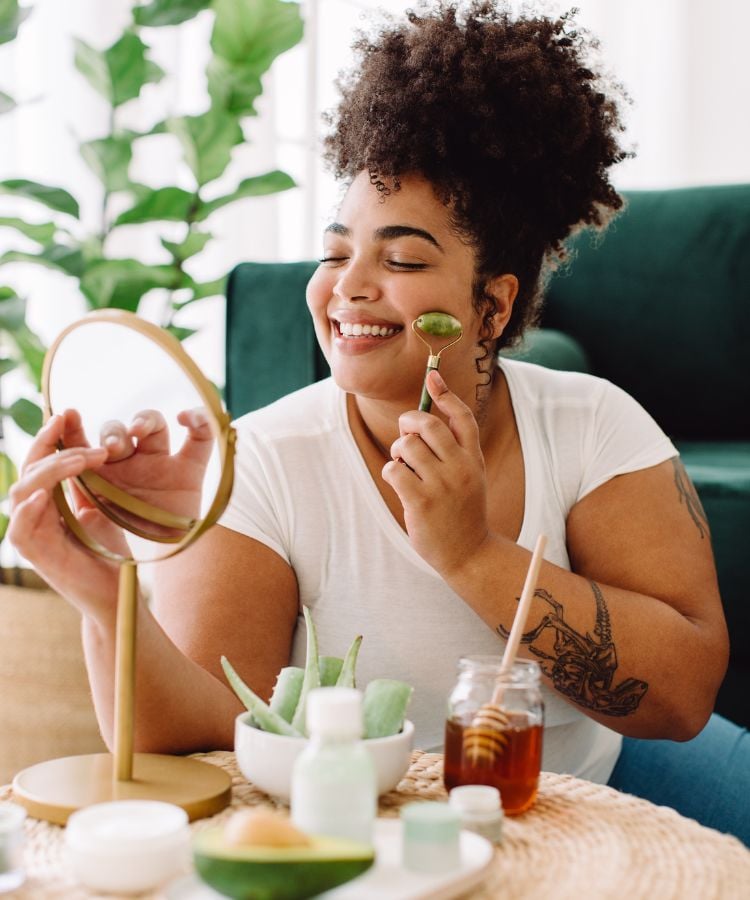 A imagem mostra uma mulher sorrindo enquanto faz uma rotina de cuidados com a pele. Ela usa um rolo de jade no rosto e segura um espelho. Na mesa, estão cremes, mel e aloe vera. O ambiente é tranquilo, com uma planta ao fundo e um sofá verde. Essa imagem representa a ideia de recompensar-se com um dia de auto cuidado, para abandonar um mau hábito.