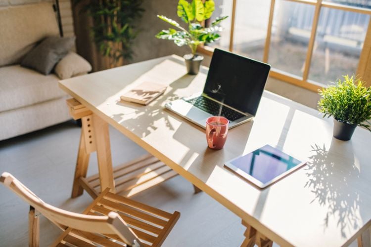 Uma mesa de trabalho minimalista é iluminada pela luz do sol que entra por uma grande janela. Sobre a mesa, há um laptop aberto, um tablet e uma caneca vermelha. Duas plantas adicionam um toque de natureza. Ao fundo, um sofá bege complementa o ambiente aconchegante e organizado.