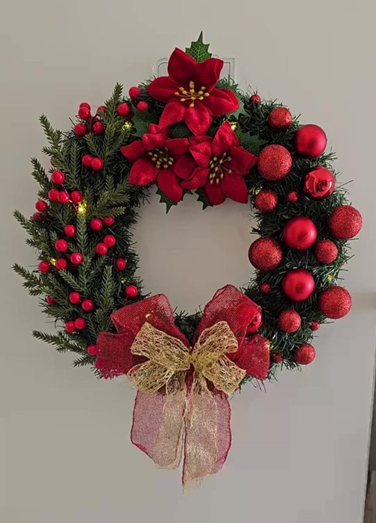 Enfeite de Natal de porta feito com ramos verdes. Decorado com flores vermelhas, bagas vermelhas e bolas natalinas. Destaca-se o laço dourado e vermelho na base, criando um visual festivo e acolhedor, ideal para celebrar a temporada natalina.