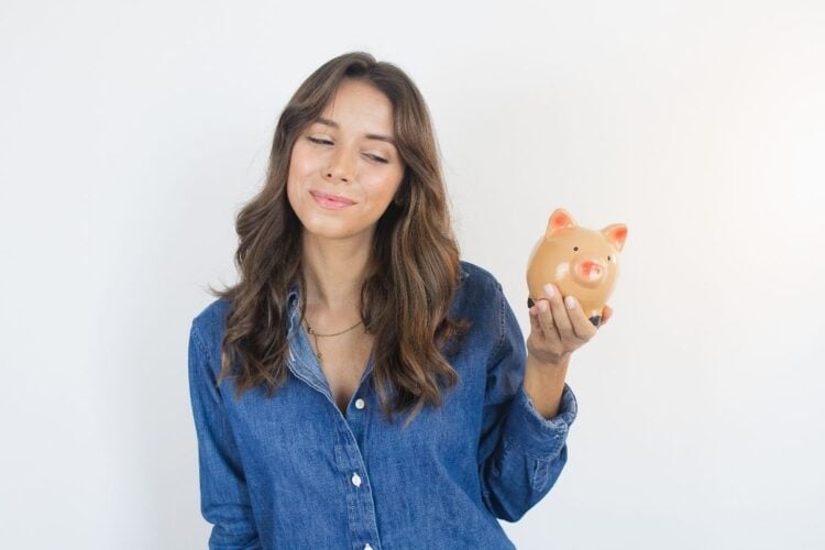 Foto de mulher morena de cabelos longos usando camisa jeans e segurando cofre de porquinho, simbolizando como investir com pouco dinheiro