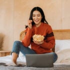 A imagem mostra uma mulher sorridente, sentada confortavelmente em uma cama, vestindo um suéter laranja e jeans. Ela está assistindo algo em um laptop e segurando uma tigela de pipoca. O ambiente é acolhedor, com decoração simples e plantas ao fundo, transmitindo uma sensação de relaxamento.