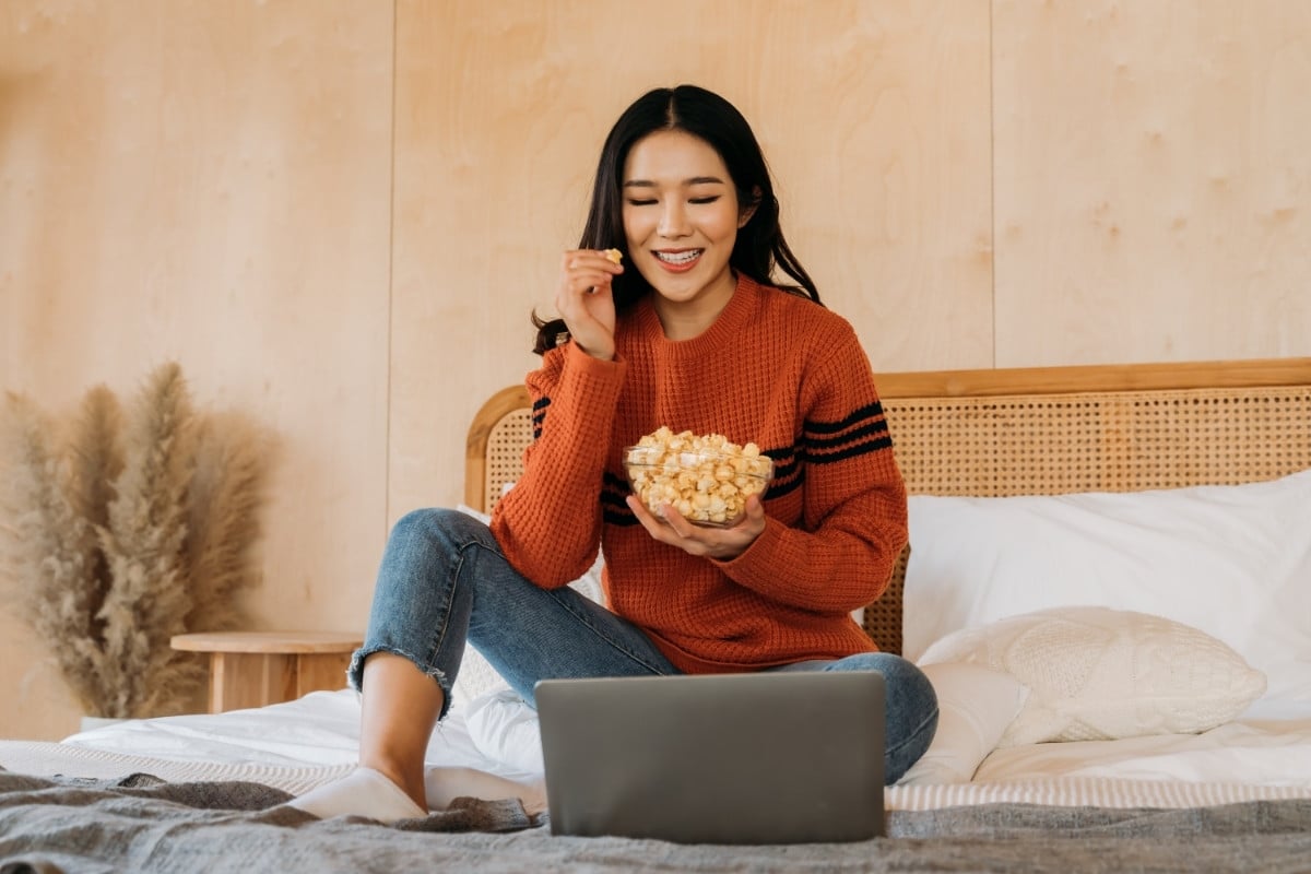 A imagem mostra uma mulher sorridente, sentada confortavelmente em uma cama, vestindo um suéter laranja e jeans. Ela está assistindo algo em um laptop e segurando uma tigela de pipoca. O ambiente é acolhedor, com decoração simples e plantas ao fundo, transmitindo uma sensação de relaxamento.