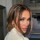 Jennifer Lopez com cabelo castanho curto e maquiagem sofisticada olha para trás, destacando seus olhos expressivos. Ela usa uma blusa de gola alta e brincos chamativos. O ambiente ao fundo é uma sala elegante, com decoração minimalista e flores brancas.