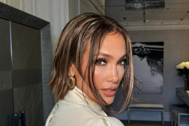 Jennifer Lopez com cabelo castanho curto e maquiagem sofisticada olha para trás, destacando seus olhos expressivos. Ela usa uma blusa de gola alta e brincos chamativos. O ambiente ao fundo é uma sala elegante, com decoração minimalista e flores brancas.