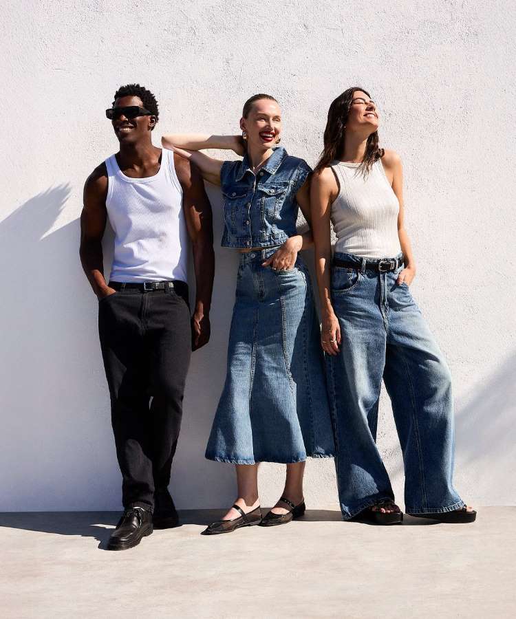 Três pessoas estão encostadas em uma parede branca, sorrindo. Uma veste camiseta branca e calça preta, outra jaqueta jeans e saia longa, e a terceira usa blusa regata branca e jeans largos. O clima é descontraído e ensolarado.