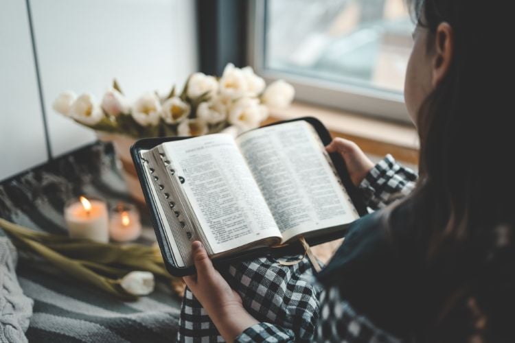 Uma mulher lê uma Bíblia junto à janela, cercada por flores brancas e velas acesas. O ambiente é tranquilo e acolhedor, iluminado por luz suave. A cena transmite paz e reflexão, com foco na leitura espiritual.