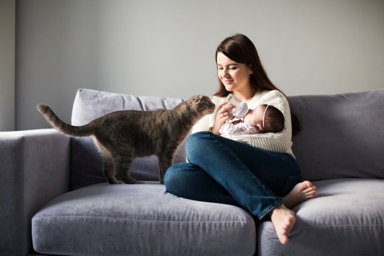 Mulher de pele clara no sofá amamentando bebê com gato ao lado 