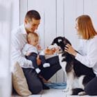 Homem de pele clara usando roupas claras, segurando bebê e mulher de pele clara com camisa branca e calça preta segurando cachorro branco e preto