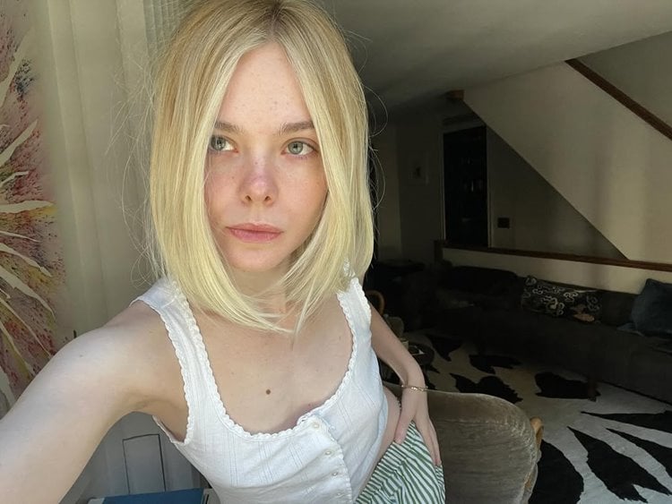 A imagem mostra Elle Fanning de cabelos loiros lisos com corte bob, usando uma blusa branca com botões. Ela está em um ambiente interno iluminado, com uma sala ao fundo que tem um sofá cinza e um tapete estampado.