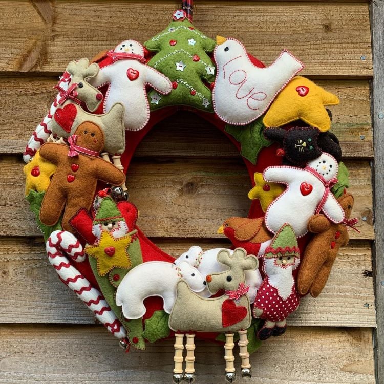 Guirlanda natalina de feltro com figuras festivas: boneco de neve, árvore de Natal, pássaro, rena, boneco de gengibre, Papai Noel e estrela. As cores vibrantes incluem vermelho, verde, amarelo e branco, com detalhes bordados. O fundo de madeira realça o artesanato acolhedor e alegre, perfeito para decoração de Natal.