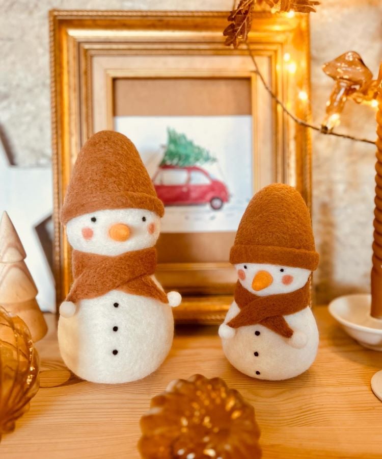 Dois bonecos de neve de feltro, com chapéus e cachecóis marrons, estão sobre uma mesa de madeira. Ao fundo, uma moldura dourada com uma pintura de carro vermelho carregando uma árvore de Natal. Luzes douradas decoram o ambiente, criando uma atmosfera acolhedora e festiva.