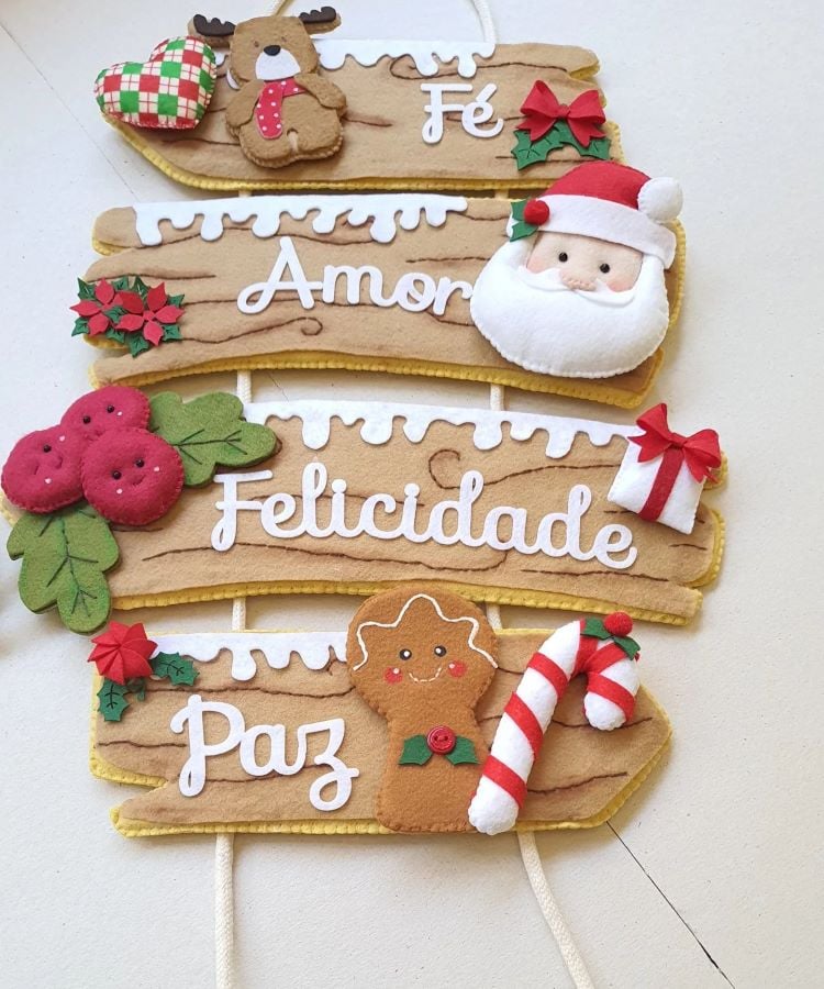 Enfeites de feltro retangulares com palavras "Fé", "Amor", "Felicidade" e "Paz". Cada um possui decoração natalina: rena e coração, Papai Noel, frutas vermelhas, boneco de gengibre e bengala doce. Os temas e cores vibrantes criam uma atmosfera alegre e festiva. Ideal para decoração de Natal artesanal.