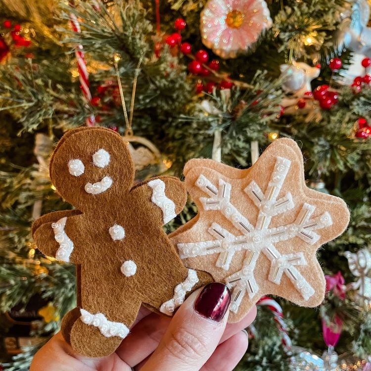 Dois enfeites de feltro são mostrados em frente a uma árvore de Natal decorada. Um é um boneco de gengibre sorridente, e o outro é um floco de neve bege com detalhes brancos. Ambos têm acabamento com bordados delicados. As luzes da árvore e enfeites vermelhos criam um fundo festivo.