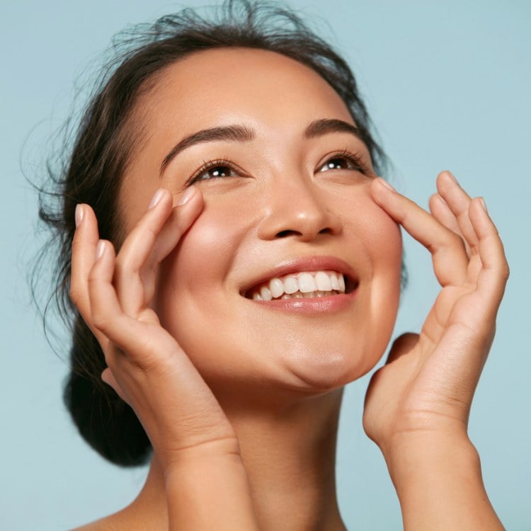 Mulher de origem asiática sorrindo, colocando as mãos no rosto, que não possui manchas nem marcas