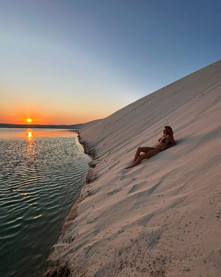 Em um cenário deslumbrante, Gisele está relaxando em uma duna de areia ao entardecer. O sol se põe no horizonte, refletindo nas águas tranquilas ao lado. A paisagem serena envolve tudo com tons quentes e suaves, criando uma atmosfera de paz e contemplação.