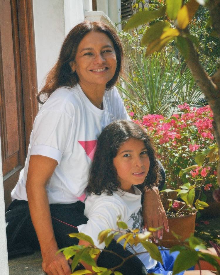 Na imagem, Dira Paes está ajoelhada ao lado de uma criança em um jardim florido. Ambas sorriem, rodeadas por flores vibrantes e folhagens exuberantes. Como uma das famosas que ficaram grávidas depois dos 40, ela demonstra alegria e serenidade, aproveitando um momento especial de conexão maternal e harmonia com a natureza.