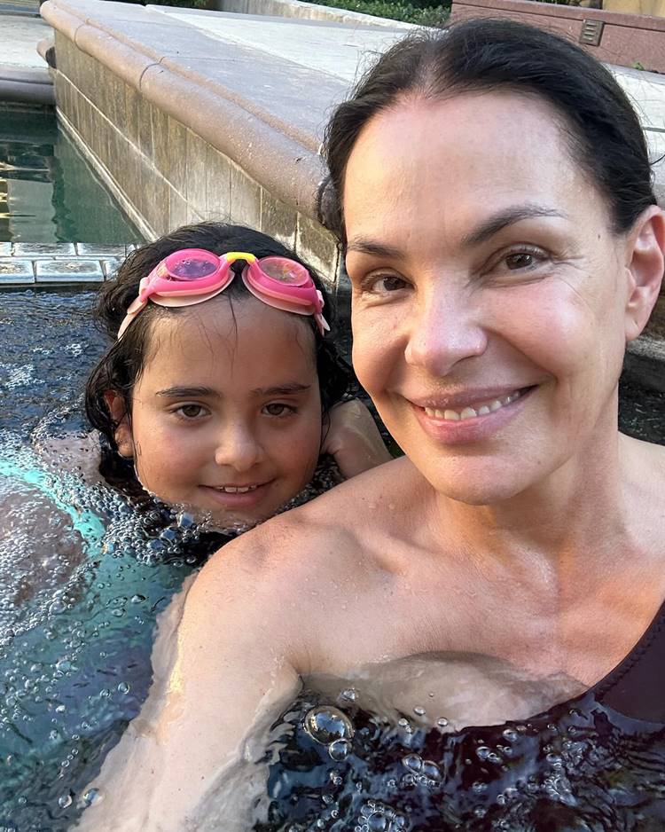 Na imagem, Carolina Ferraz está sorrindo ao lado de uma criança em uma piscina. Ambas se divertem na água, com a criança usando óculos de natação rosa. Como uma das famosas que ficaram grávidas depois dos 40, ela exala felicidade e aproveita um momento de lazer tranquilamente.