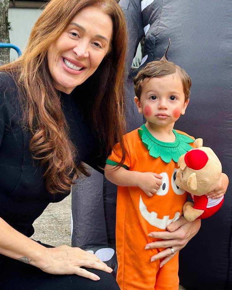 Na imagem, Claudia Raiaestá ajoelhada ao lado do seu filho vestido como abóbora, segurando um brinquedo. Ela sorri calorosamente, criando uma cena encantadora. Como uma das famosas que ficaram grávidas depois dos 40, ela irradia alegria maternal. O cenário é descontraído e festivo, capturando um momento feliz.