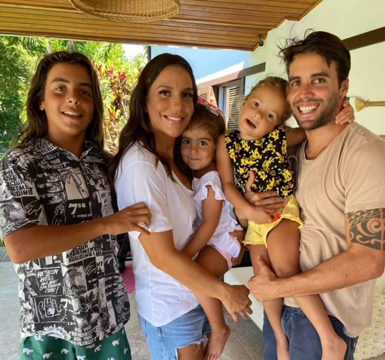 Nesta imagem, Ivete Sangalo está cercada por sua família em um ambiente doméstico acolhedor. Ela sorri ao lado de um homem e três crianças. Todos aparentam estar felizes e descontraídos. O cenário ao fundo revela um espaço arejado com vegetação, criando uma atmosfera de união e harmonia familiar.