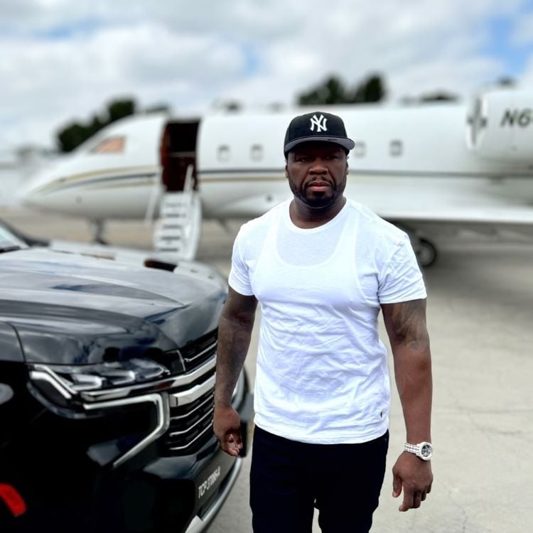 50 Cent está parado ao lado de um carro preto, em frente a um jato particular. Ele usa boné e camiseta branca. A imagem evoca luxo e poder, ele é um dos famosos que apoiam Trump ou Kamala, indicando a complexidade e diversidade de opiniões no mundo das celebridades.