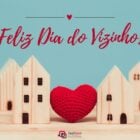 Foto de casas de madeira ligadas por um coração de crochê. Acima, lemos "Feliz Dia do Vizinho"