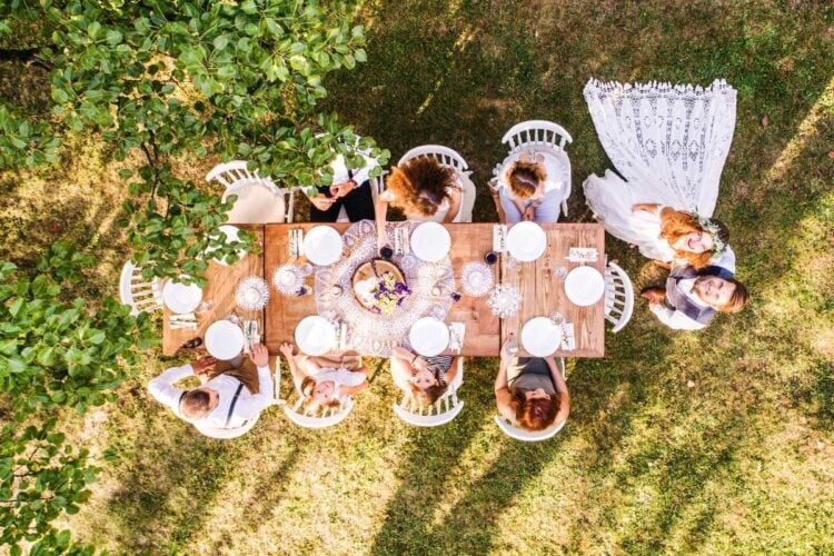 Uma mesa rústica ao ar livre vista de cima, cercada por pessoas sorridentes, em um ambiente natural com árvores. O casal de noivos está de pé, felizes, ao lado da mesa, sugerindo uma celebração íntima e harmoniosa.