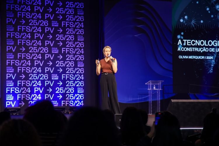 Olivia Melquior usando blusa marrom e calça preta falando ao microfone no palco do Focus Fashion Summit 