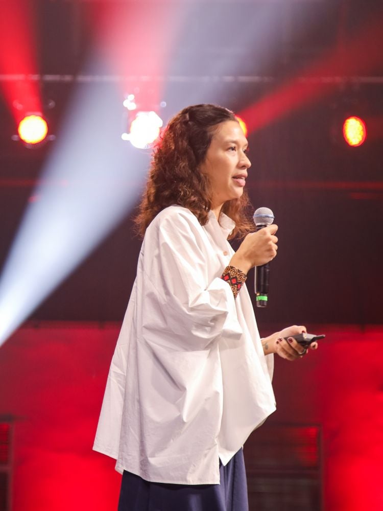 Jaqueline Amaral usando blusa e falando ao microfone, com jogo de luzes brancas e vermelhas ao fundo 