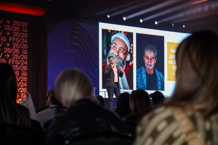 Marcos Bedendo usando terno no palco do evento, com fotos de homens no telão atrás dele 