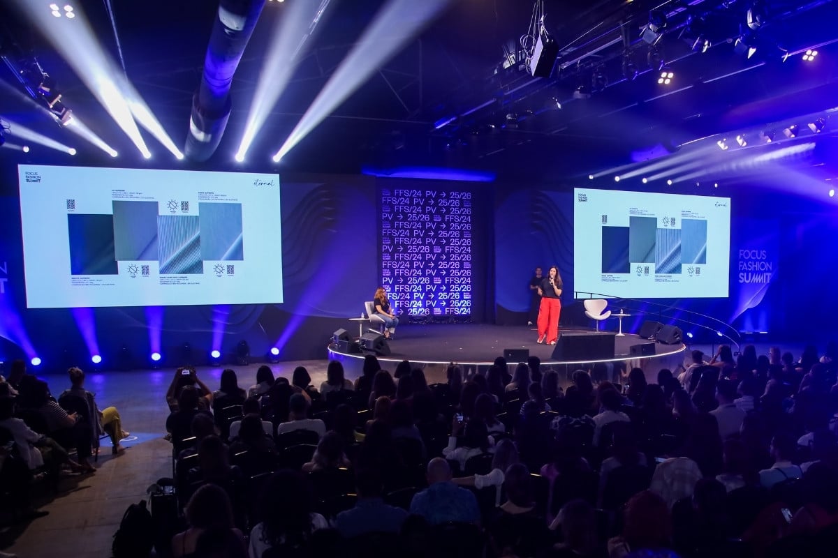 Foto de pessoa palestrando para a plateia em palco do Focus Fashion Summit, com telões com gráficos